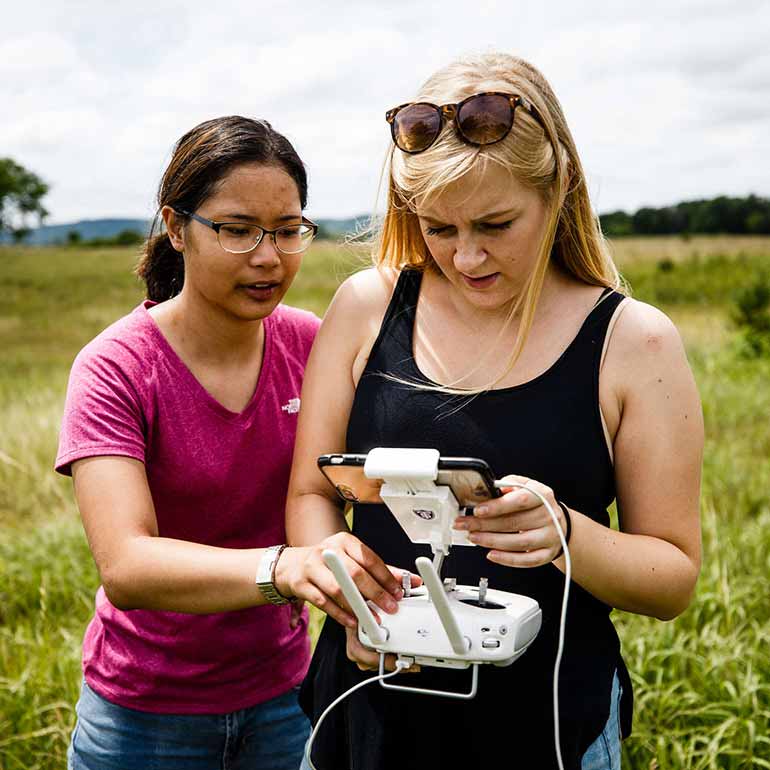 Two students stand in a prairie while flying a drone.