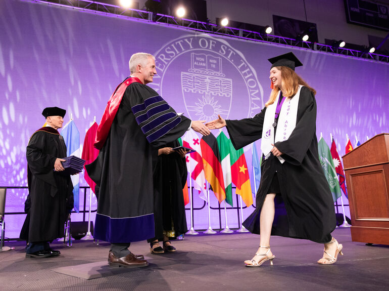 A student in cap and gown walks across the graduation commencement stage to shake President Vischer’s hand