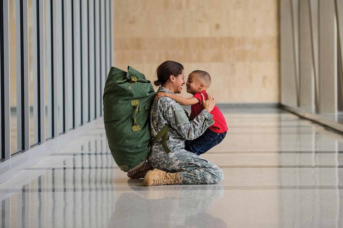 Female dressed in fatigues kneels on ground to greet child.