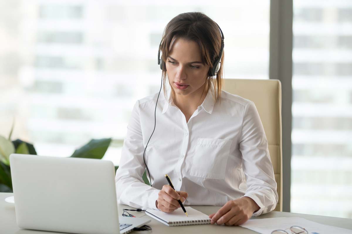 A woman wearing headphones sits at a laptop taking notes.