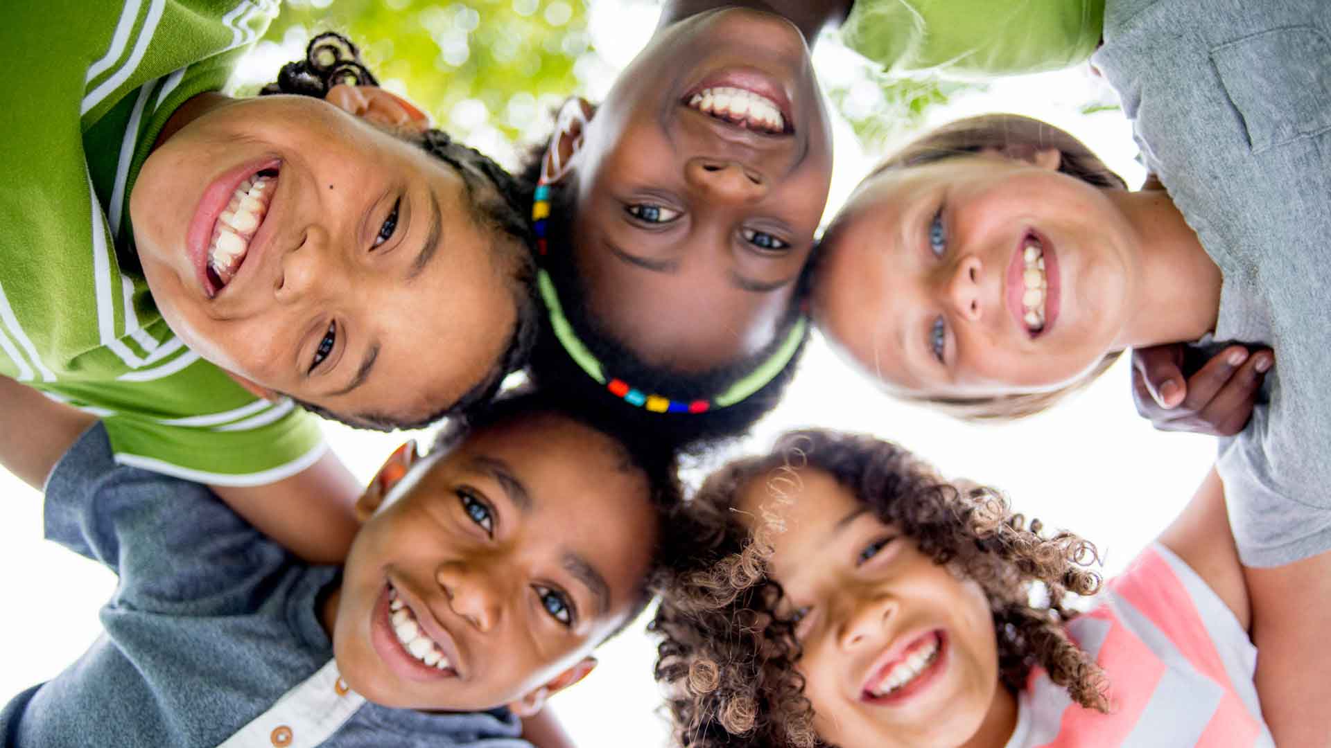 Upward view of five children from different ethnicities in a circle smiling for the camera.