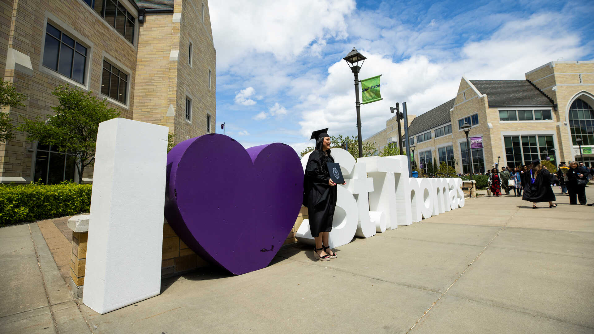 Student in graduation cap and gown standing in front of sign saying I love St. Thomas