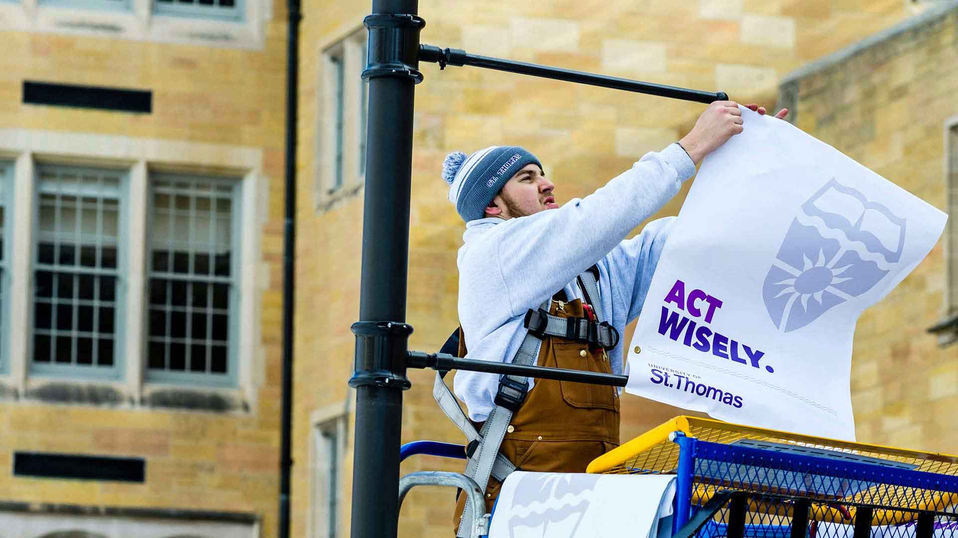 Student hangs a sign reading "Act Wisely" on a lamp post. 