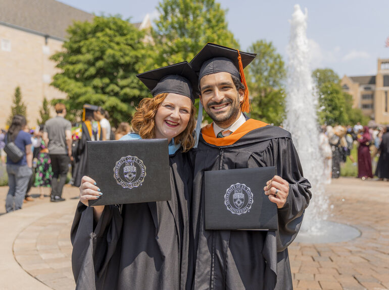 Two graduate students outside by a fountain, holding up their diplomas after their graduation ceremony.