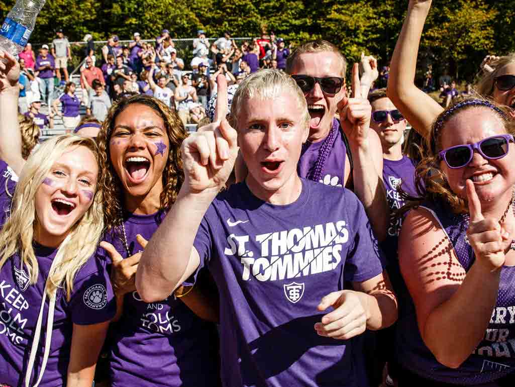 A group of students wearing purple St Thomas Tommies shirts cheer for the camera