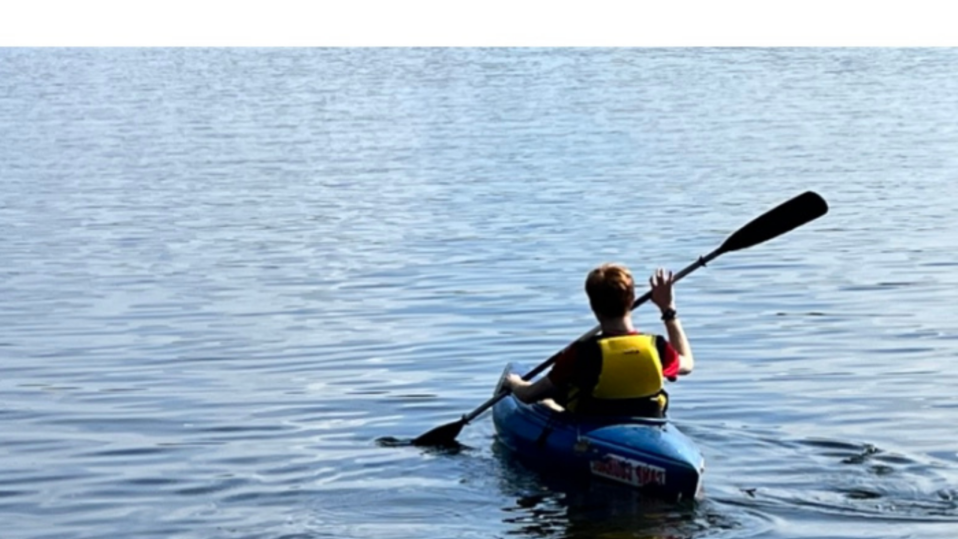 A person wearing a yellow life vest paddles a blue kayak alone on a calm lake, heading toward the open water under a clear sky. The peaceful scene suggests reflection and solitude, likely part of a retreat experience.