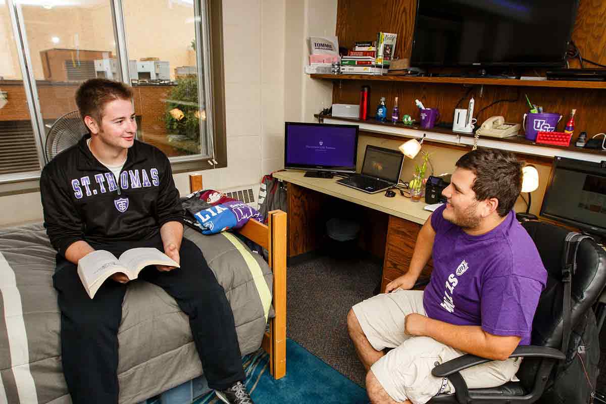 Student Anthony Hughes sits on a bed with a book while talking with friend Austin Maffei in a dorm room.
