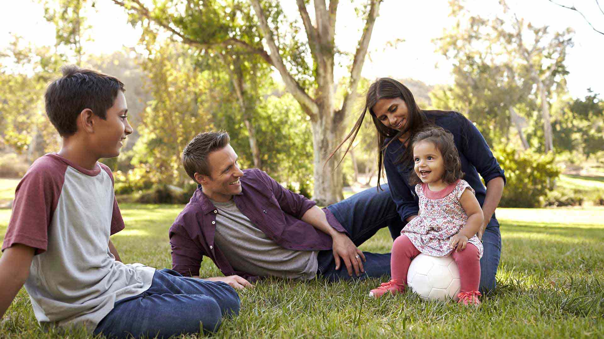 A family of four sits on the grass, the youngest child sits on a ball and smiles at her brother.