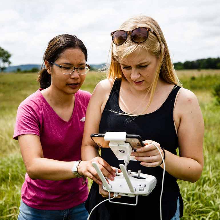 Two students stand in a prairie while flying a drone.