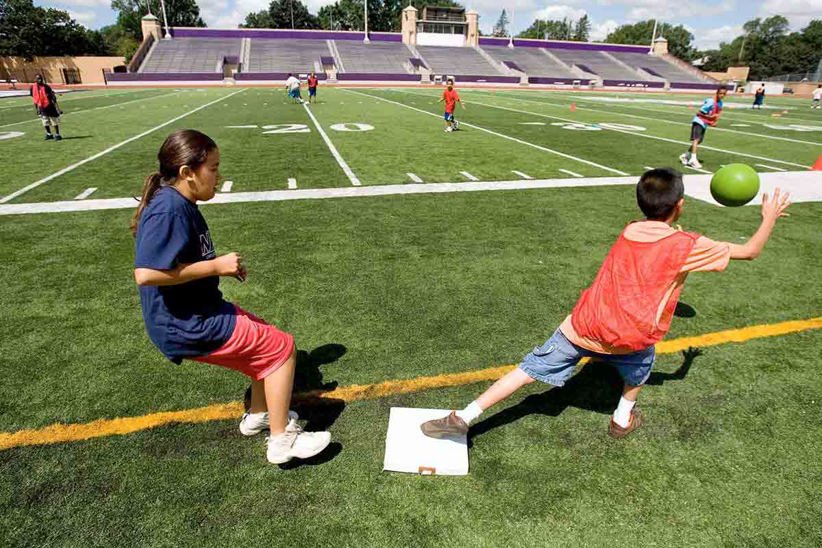Children play kickball on football field.