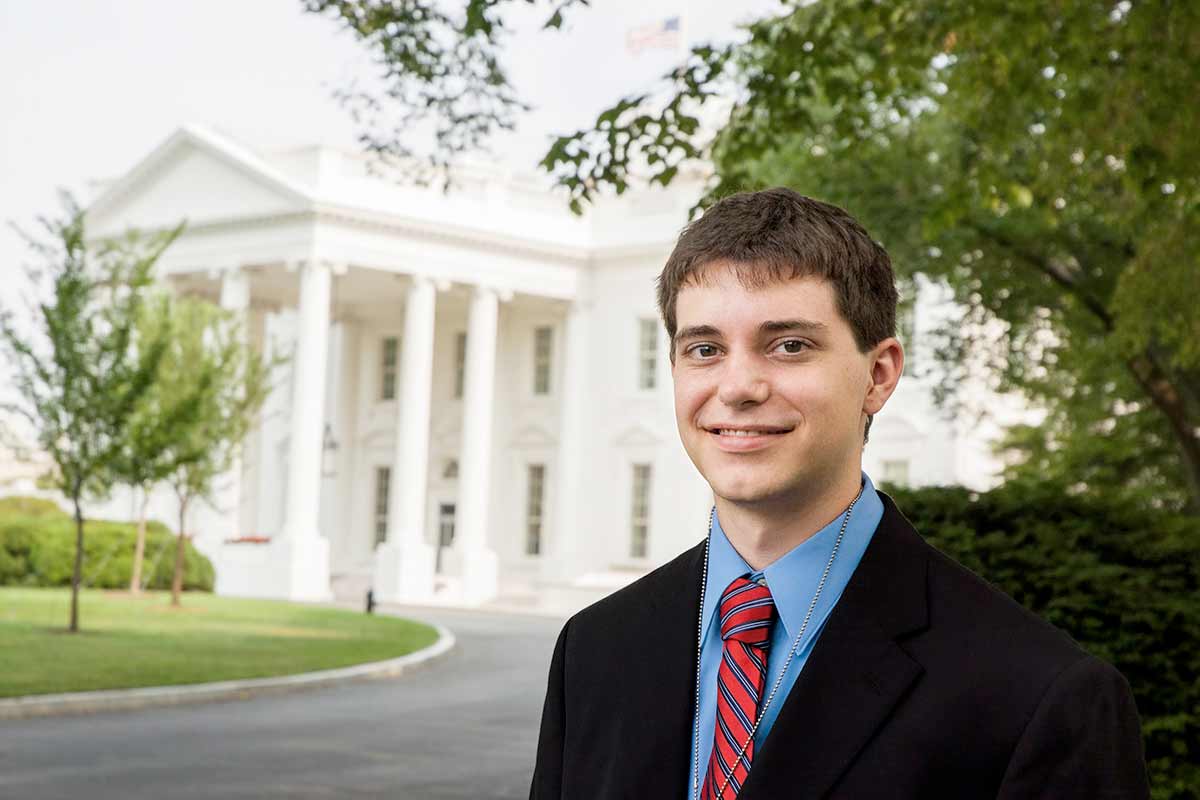 An individual in a suit poses in front of the White House.