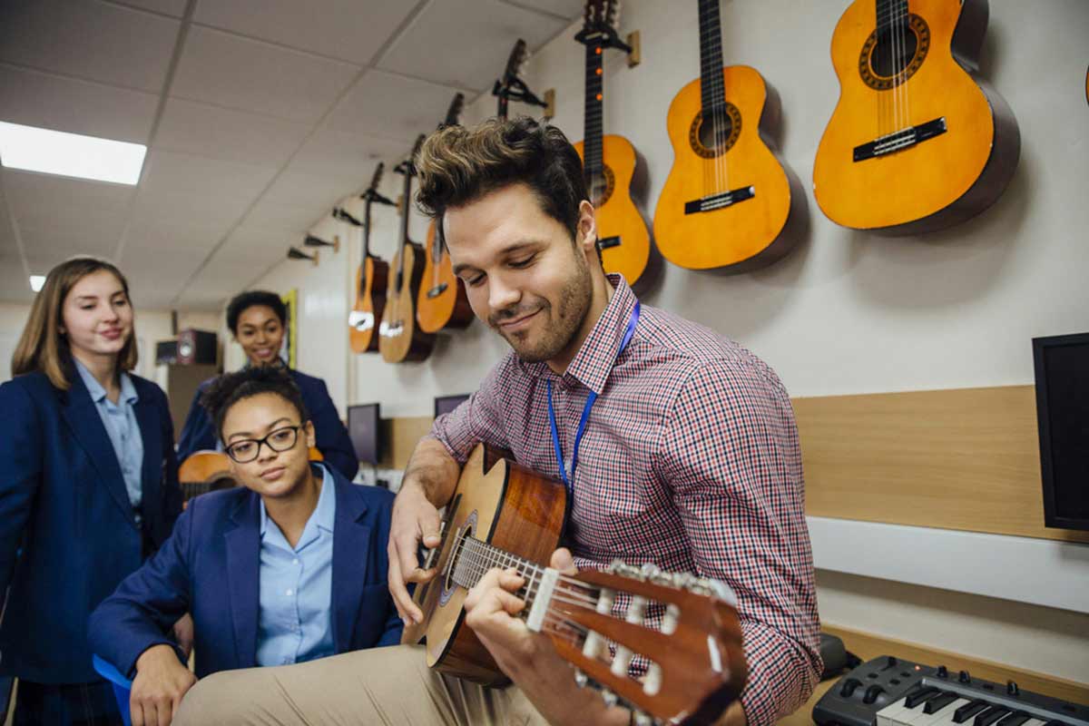 An individual is playing guitar while others look on in the background.