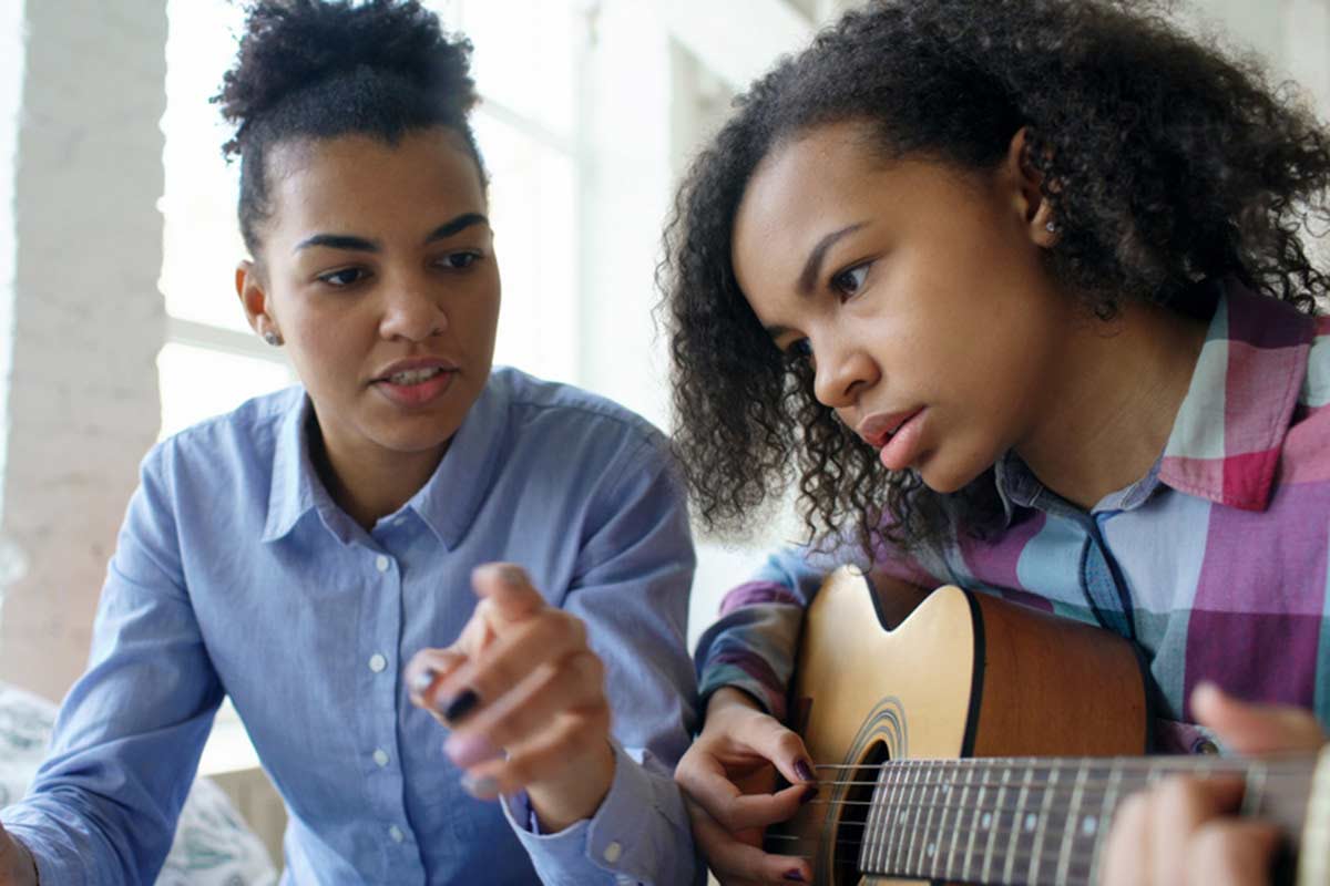 Two people sit side-by-side while playing guitar.