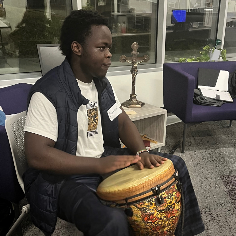 A Black male student wearing a navy vest and white t-shirt plays a colorful djembe drum. Behind him sits a crucifix on a small table and an open binder on a purple chair, suggesting a spiritually rooted gathering space.