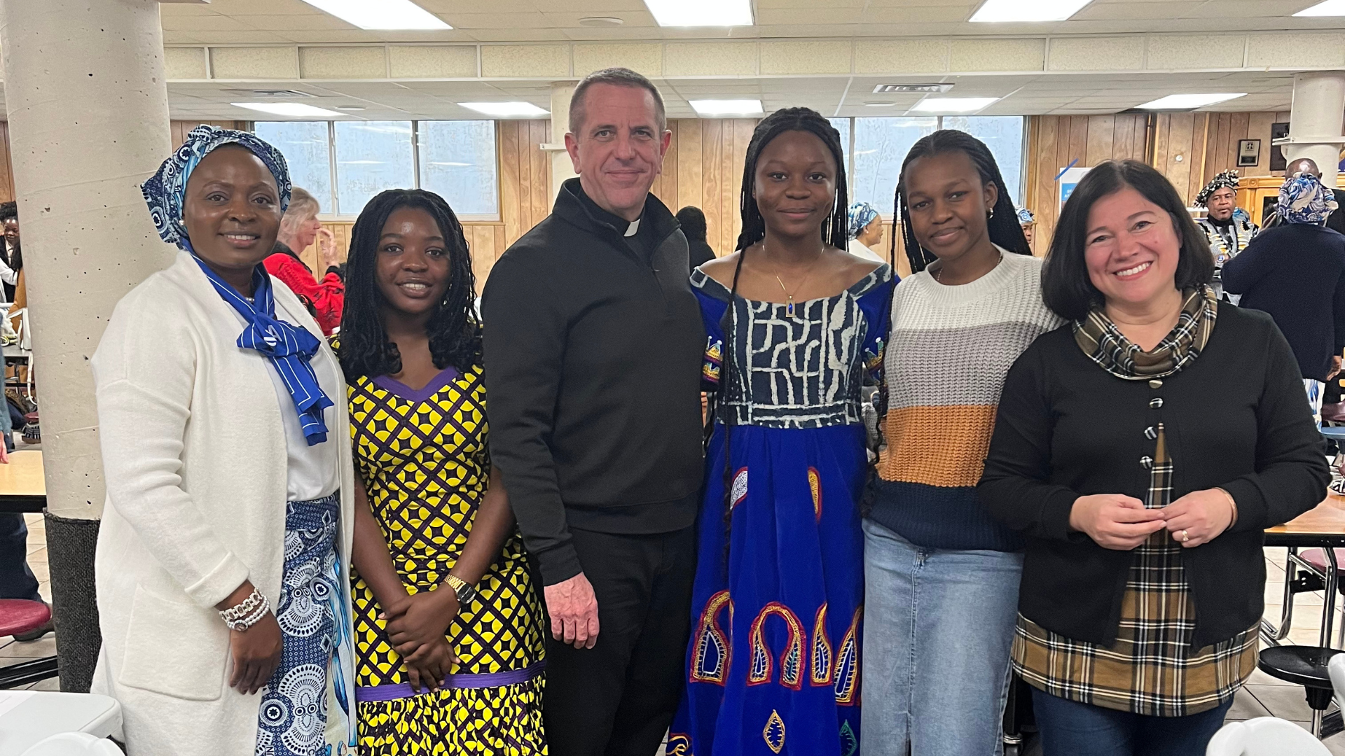 Six people stand together and smile for the camera, including a Catholic priest and five women dressed in colorful, culturally significant attire. They are gathered in a community hall, likely during a Afro Thanksgiving.