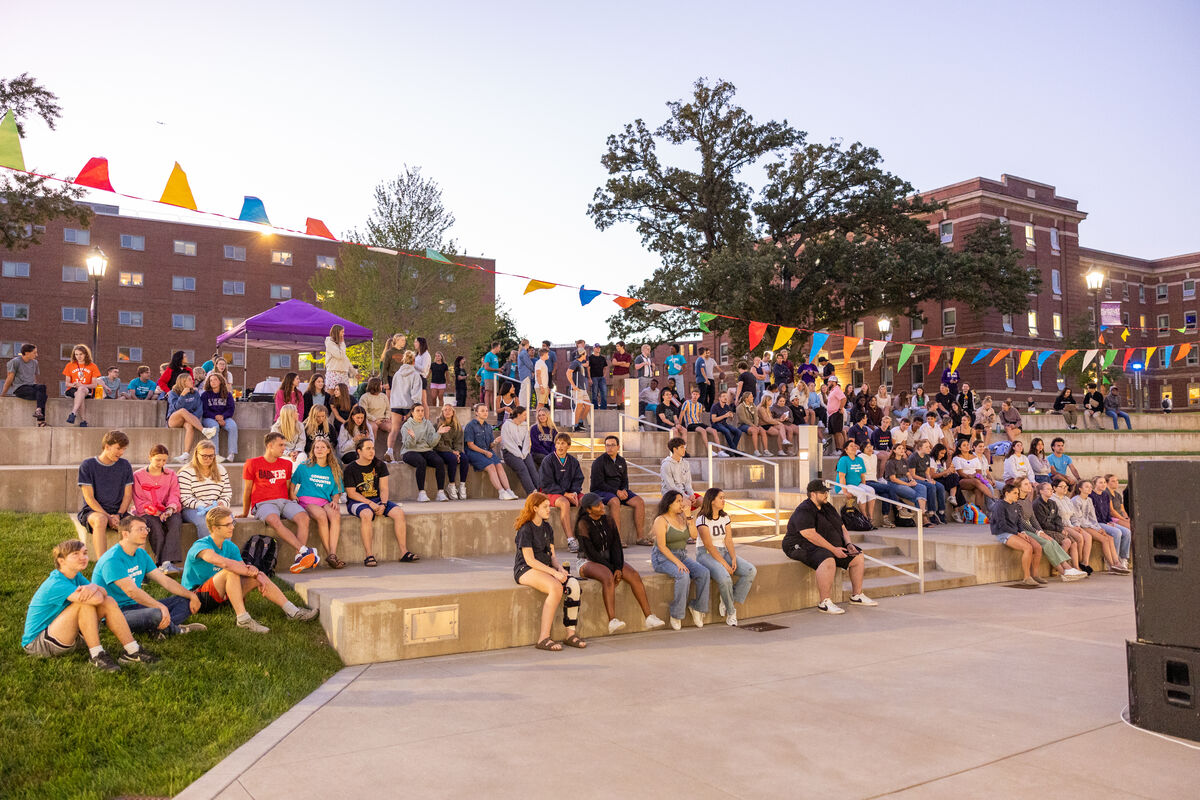 students sit outside on steps to watch a performance