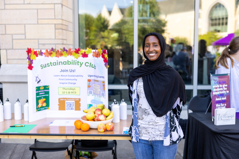 Student smiling in front of a Sustainabilty Club tri-fold poster.